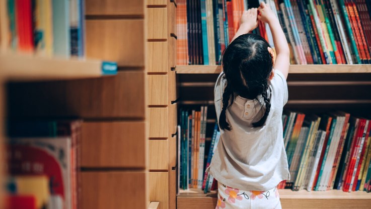 Flicka sträcker sig efter en bok i en bokhylla.