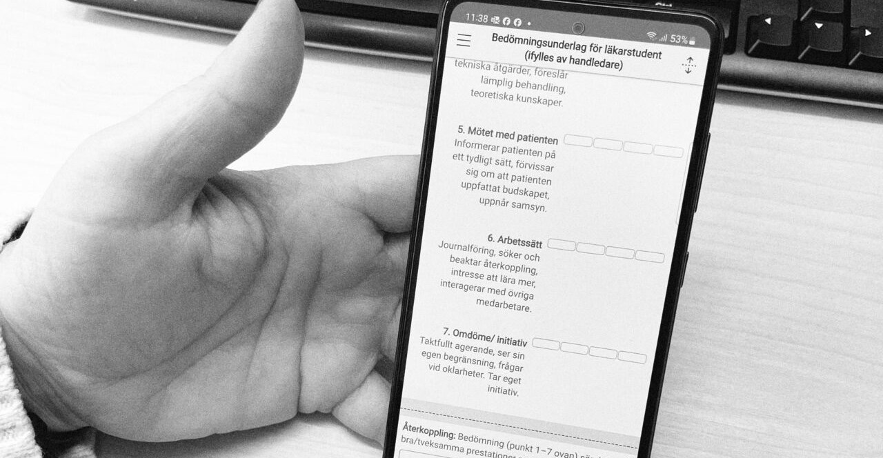En hand håller i en mobiltelefon som visar en app, i bakgrunden syns ett skrivbord och ett tangentbord.