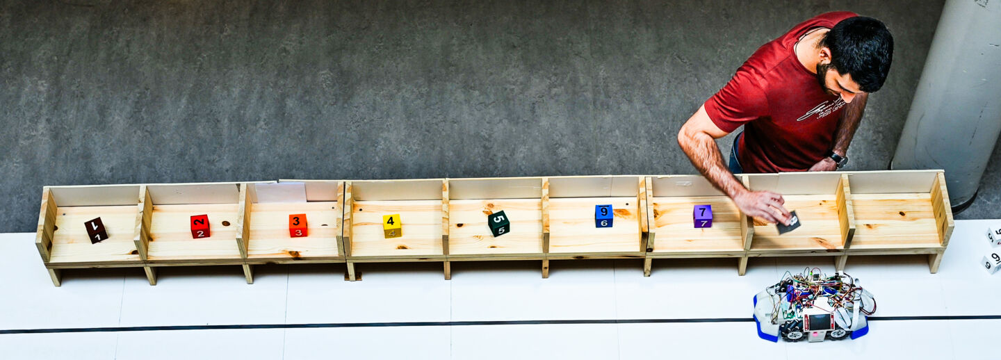 En ung man sätter klossar med olika siffror i träfack, i förgrunden kör en robot