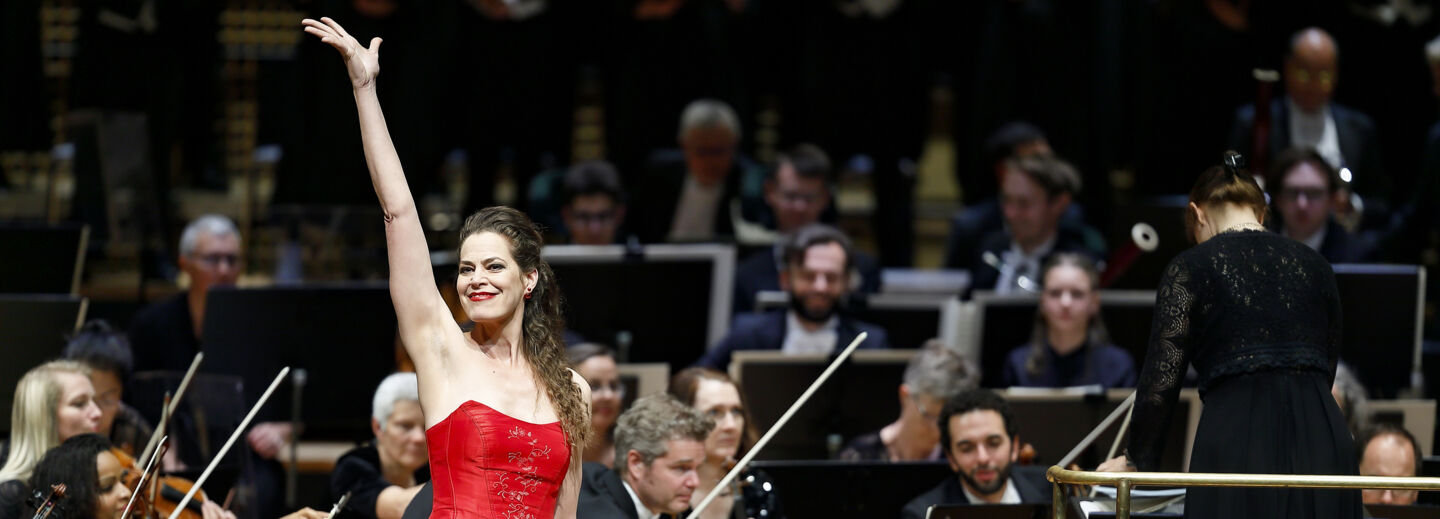 Happy female singer in a red dress holds up one arm in the air in front of an orchestra