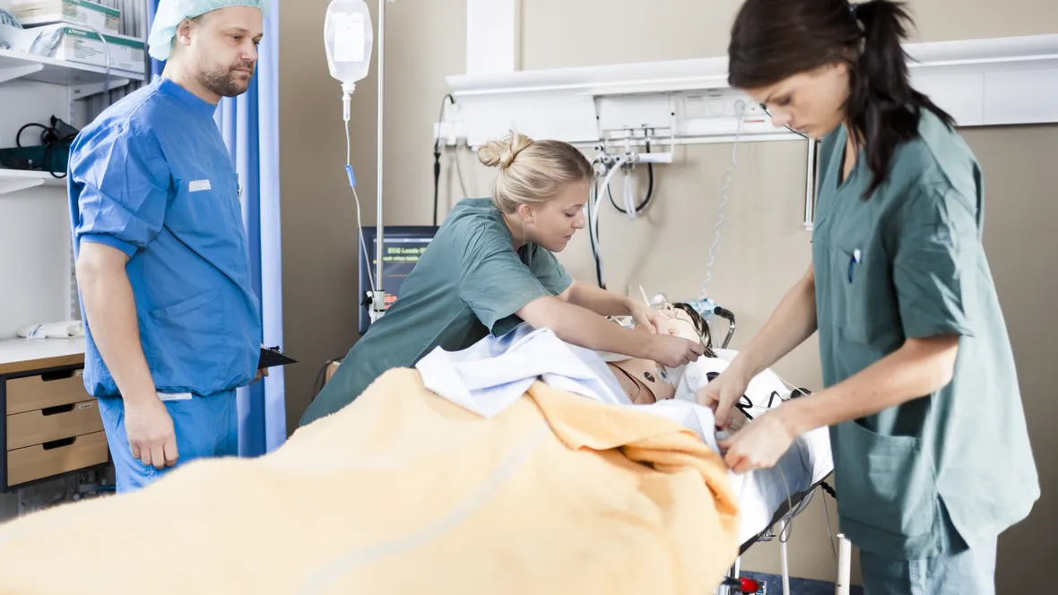 Three people in hospital clothes practice practical skills on a manikin lying in a hospital bed.