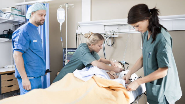 Tre personer i sjukhuskläder tränar praktiska färdigheter på en övningsdocka som ligger i en sjukhussäng.