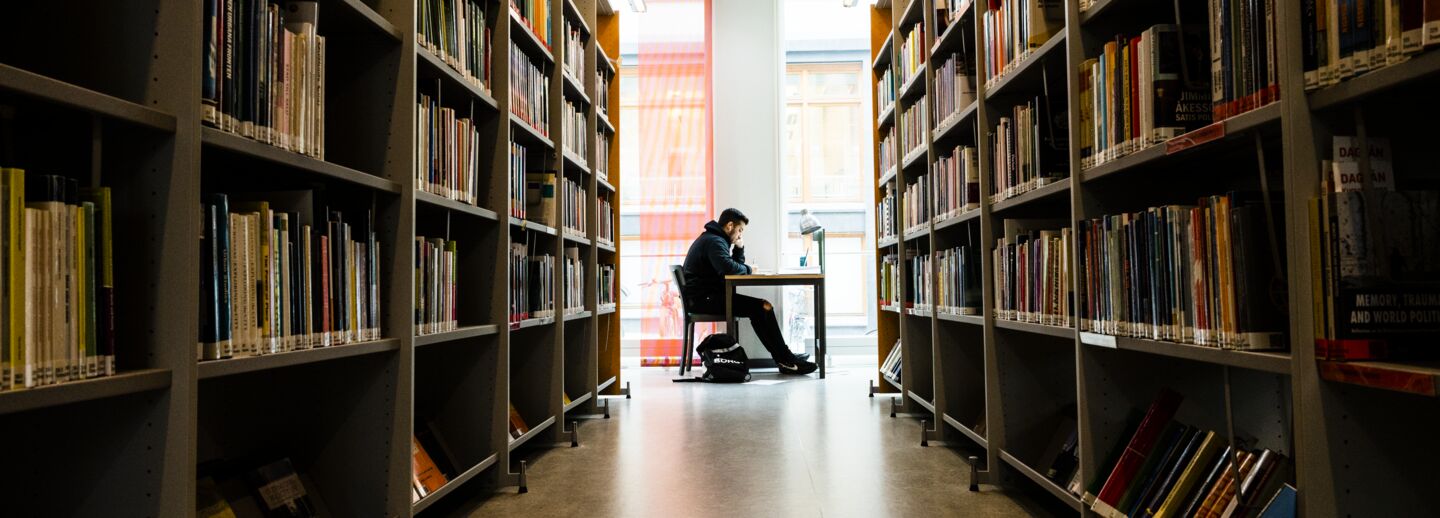 Two rows of book shelves in a library. In the space between them, a person sitting at a table in seen at a distance.