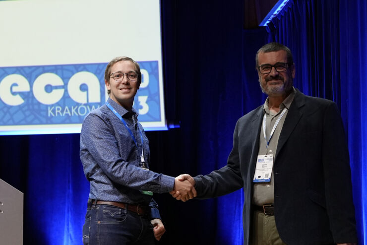 Two men at a stage shaking hand and smiling against the public, one of them holding a paper