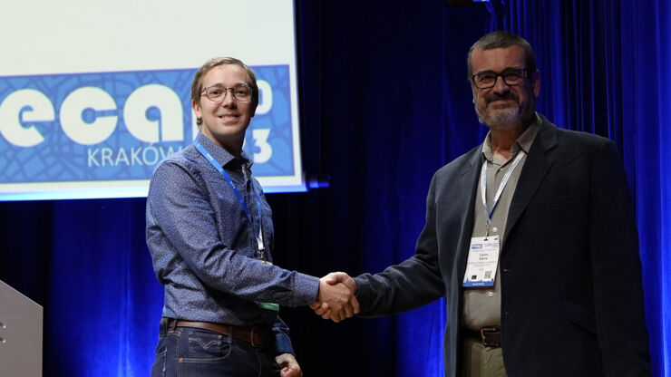 Two men at a stage shaking hand and smiling against the public, one of them holding a paper