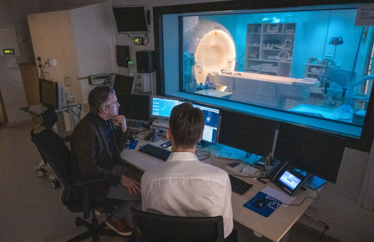 Two researchers study a computer screeen and a magnetic resonance tomograph is seen through a window pane.