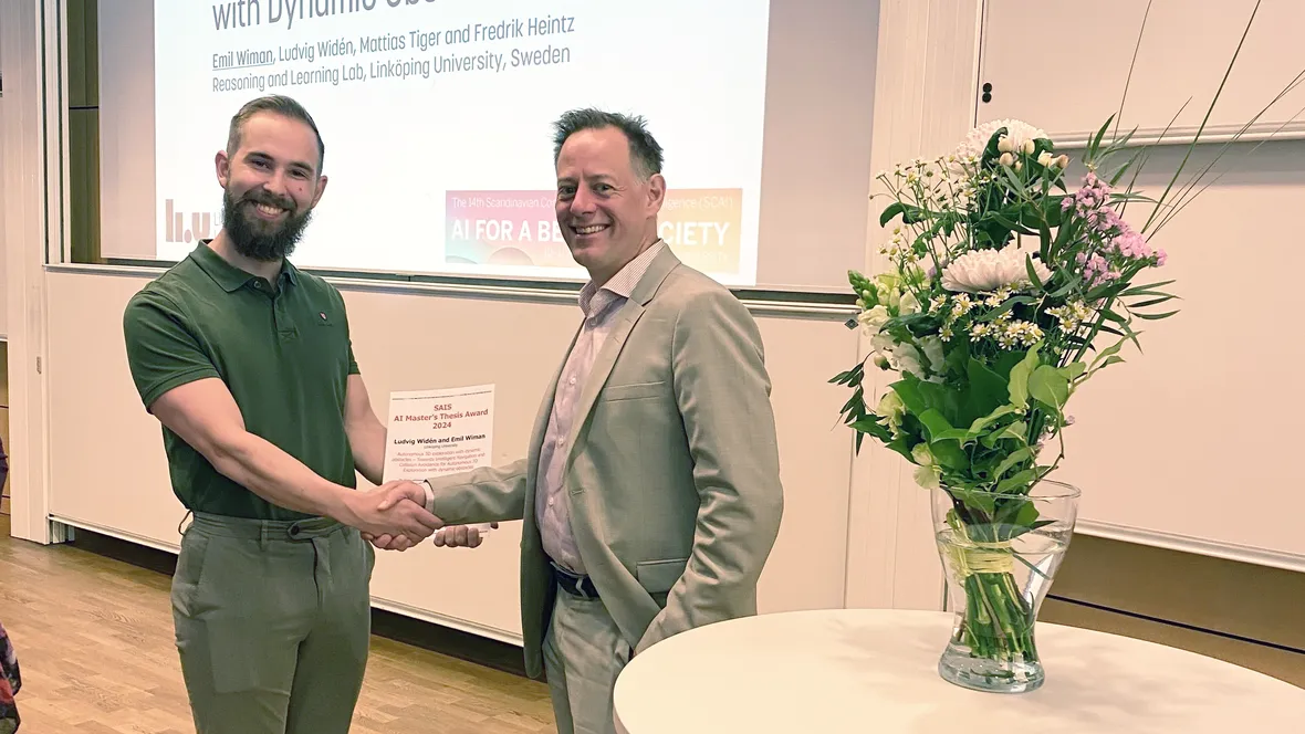 Två män längst fram i föreläsningssal tar i hand, den ena med diplom i handen