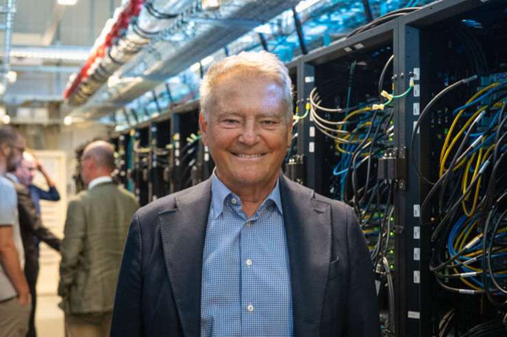 A man in a checked shirt and jacket standing in front of a supercomputer.