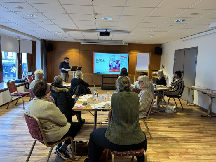 Konferensrum med kvinna som föreläser intill en storbildsskärm för en mindre publik.