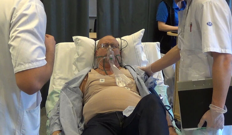 Medelålders man med syrgasmask ligger i sjukhussäng med vårdpersonal omkring sig.
