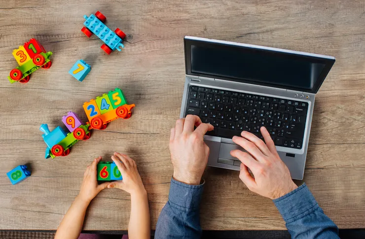 Close-up of children's hands playing with colourful blocks near an adult working on a laptop.