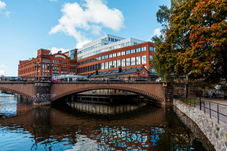 Vatten framför en bro och en byggnad. Blå himmel och ett träd i höstfärger.