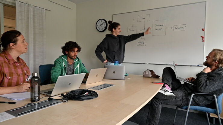  Fyra personer i ett mötesrum, tre sittande och en stående, diskuterar ett flödesschema på en whiteboard. Bärbara datorer står på bordet, och en klocka hänger på väggen.