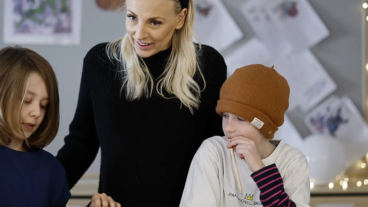 En klassrumsmiljö med en kvinna, troligen en lärare, som står bredvid två barn. Läraren, klädd i en svart tröja, interagerar med eleverna. Ett barn med axellångt hår har på sig en blå tröja och är fokuserad på en aktivitet. Det andra barnet har en brun mössa och en vit tröja med en Jean-Michel Basquiat-design, och håller en penna medan det funderar. I bakgrunden finns papper uppsatta på väggen och ljusslingor, vilket skapar en mysig och kreativ miljö.