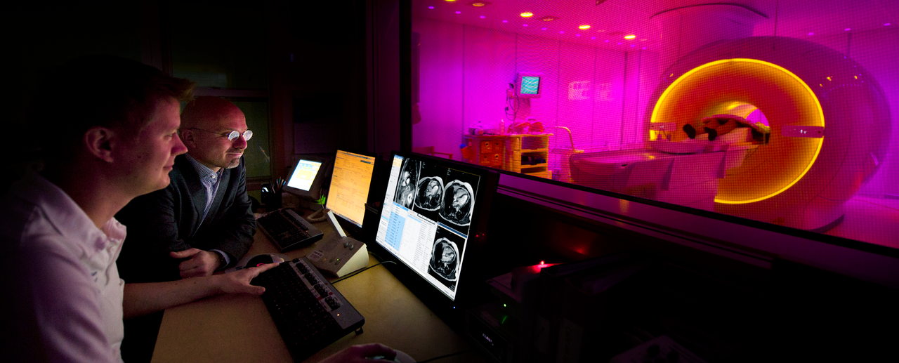 Photo of two persons in the control room in front of a MR camera