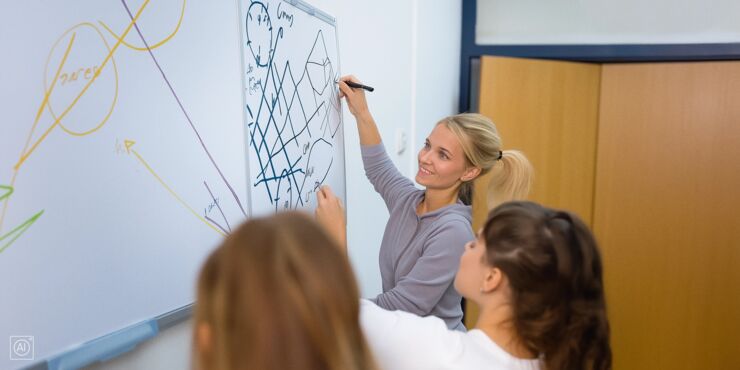 En lärare och två studenter arbetar tillsammans vid en whiteboard fylld med matematiska formler, diagram och färgglada linjer. Läraren, som ser glad och fokuserad ut, skriver eller ritar på tavlan medan studenterna hjälper till och diskuterar.