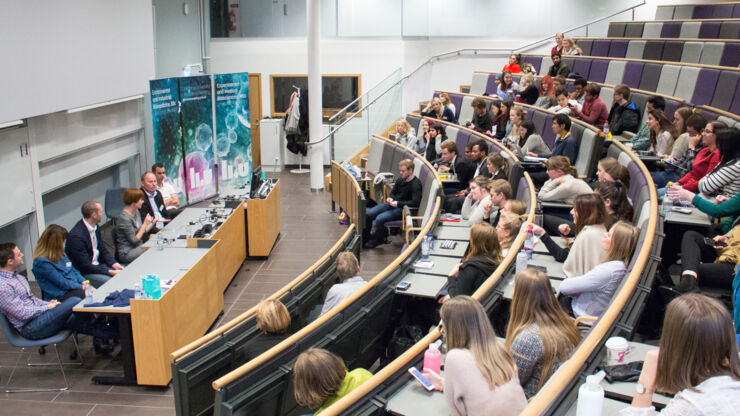 A lectur hall full of students with a panel with six participants in the front.