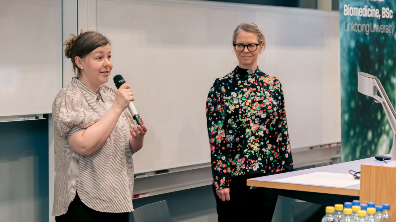 Two women in a lecture hall with a powerpoint presentation in the background.