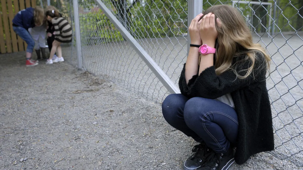 A girl is on her knees with her hands over her face; further away, some children are whispering.
