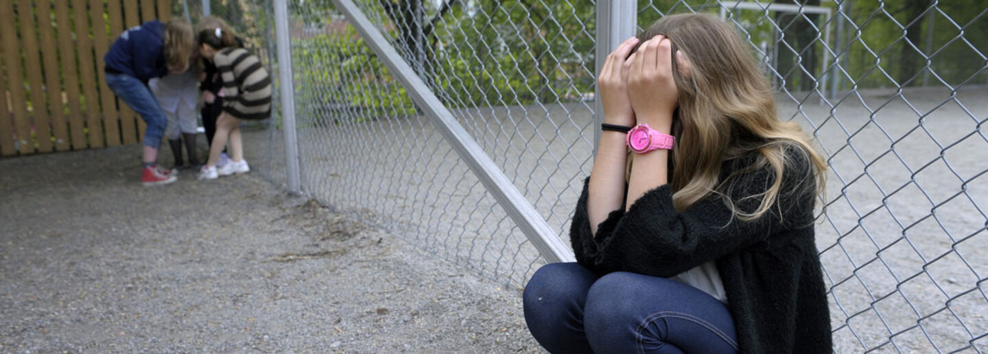 A girl is on her knees with her hands over her face; further away, some children are whispering.