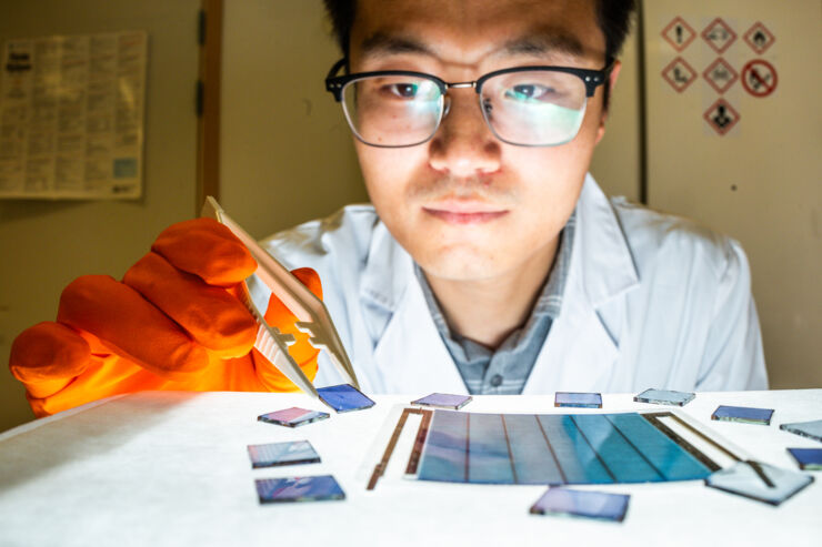 Researcher in lab coat holds blue solar cell with tweezers.