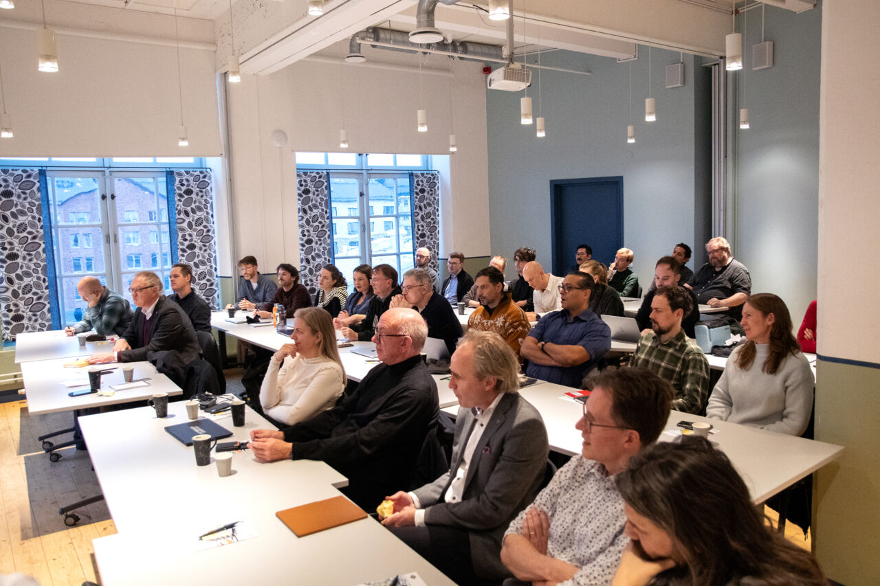 Ungefär 30 personer sitter i rader bakom skrivbord, så som i ett klassrum. 