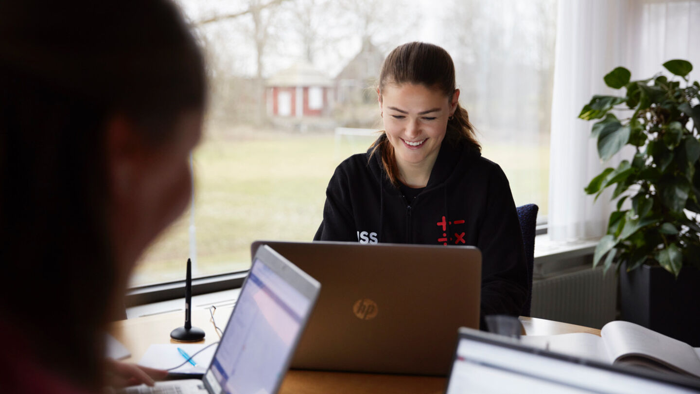 Glad ung kvinna jobbar framför dator med böcker bredvid. Har tröja med symbolen av mattecoach online